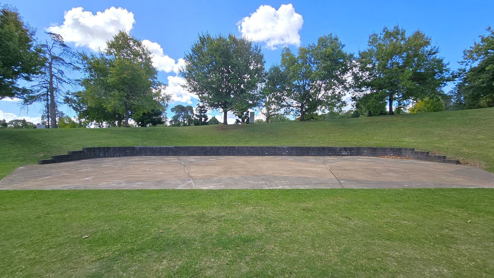 A grassy area with a concrete amphitheatre in the centre, featuring a semi-circular stage. The stage is bordered by low, dark stone walls. In the background, a row of trees lines the top of a gentle slope, with a mix of leafy green foliage. The sky is blue with scattered white clouds. The foreground consists of a well-maintained grassy lawn.