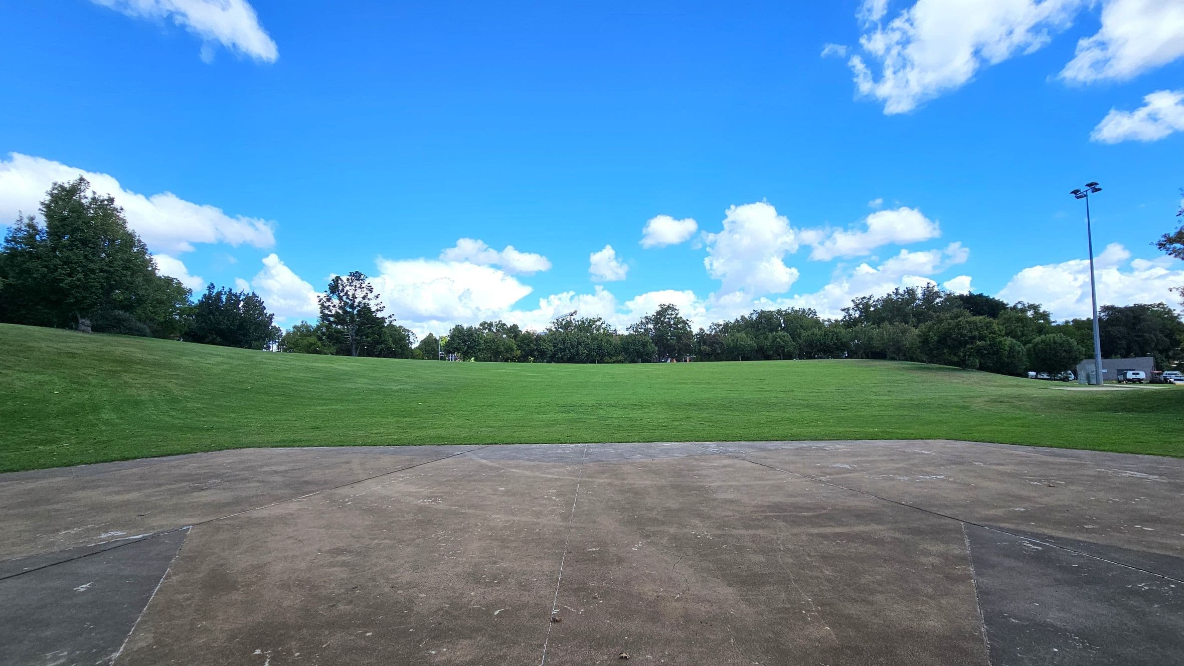 A wide expanse of green grass stretches across the middle of the image, bordered by a line of trees in the background. In the foreground, there is a concrete surface with visible wear and markings. On the right, a tall floodlight pole stands near a small building and some parked vehicles. The sky is bright blue with scattered white clouds.