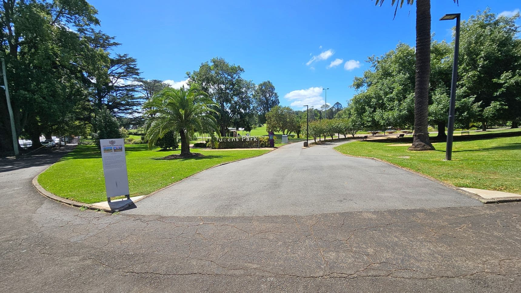 A paved path leads into a park with a sign reading "Queens Park" in the centre. On the left, there's a large tree and a sign with information. To the right, a tall palm tree and a lamp post are visible. The park is lush with green grass and various trees, including palm and deciduous types. Benches are scattered throughout the area, and the sky is clear and blue.