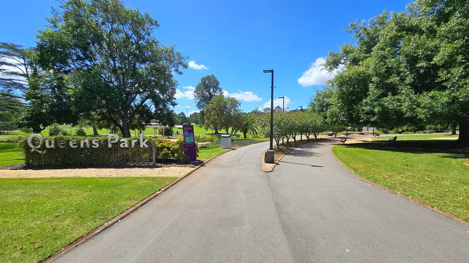 A paved path runs through a park, flanked by well-maintained grass and trees. On the left, a sign reads "Queens Park" with a large tree behind it. To the right, more trees provide shade over benches. The path is lined with street lamps and curves gently, leading deeper into the park. In the background, open grassy areas and additional trees are visible under a clear blue sky.