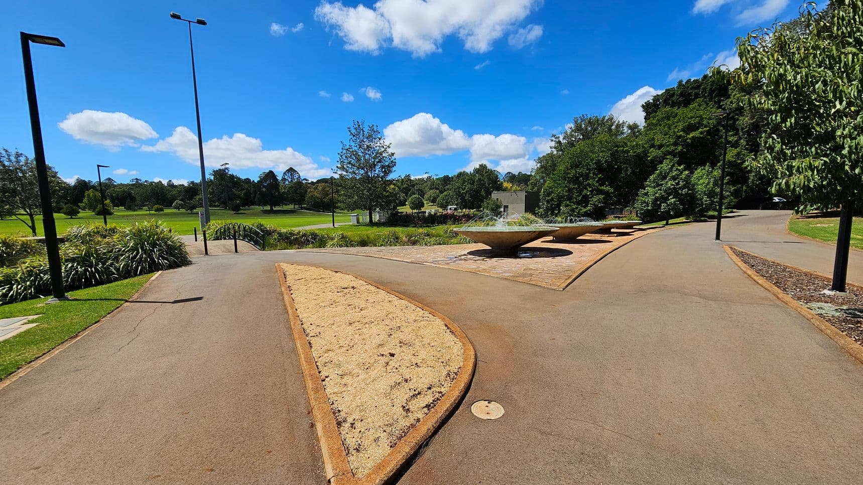 A park scene with a paved path splitting into two directions. On the left, there are tall street lamps and lush green grass with scattered trees in the distance. In the centre, a sandy patch is bordered by the path. On the right, more trees and shrubs line the path, with a series of large, shallow fountains on a stone platform. The background features a mix of open grassy areas and dense tree coverage.