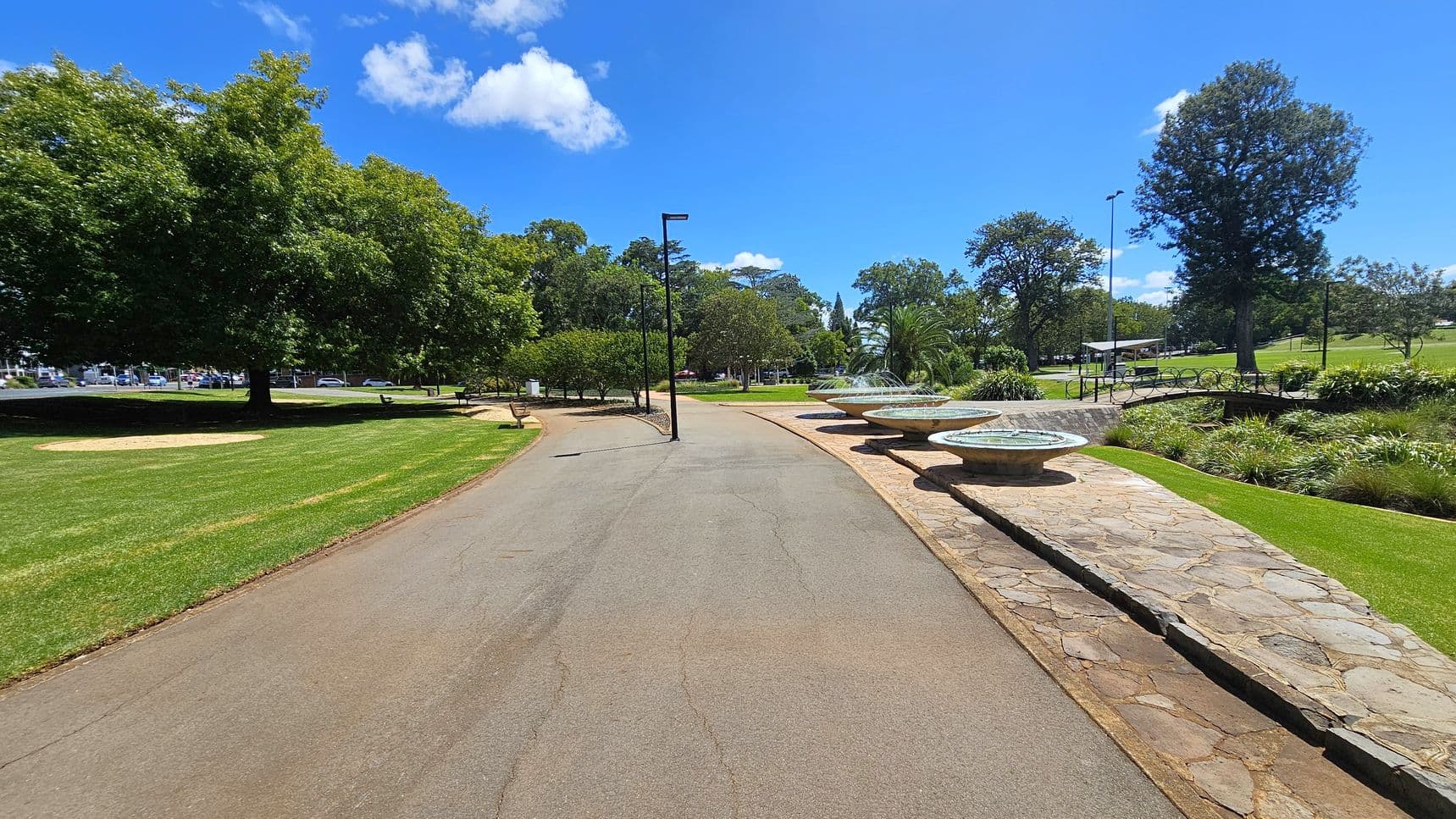 A paved pathway runs through a park, flanked by lush green grass. On the left, large trees provide shade, with a few benches visible. On the right, a series of circular fountains are set on a stone platform, surrounded by manicured plants. In the background, more trees and a small structure can be seen. A few street lamps line the path, and the sky is clear with a few clouds.