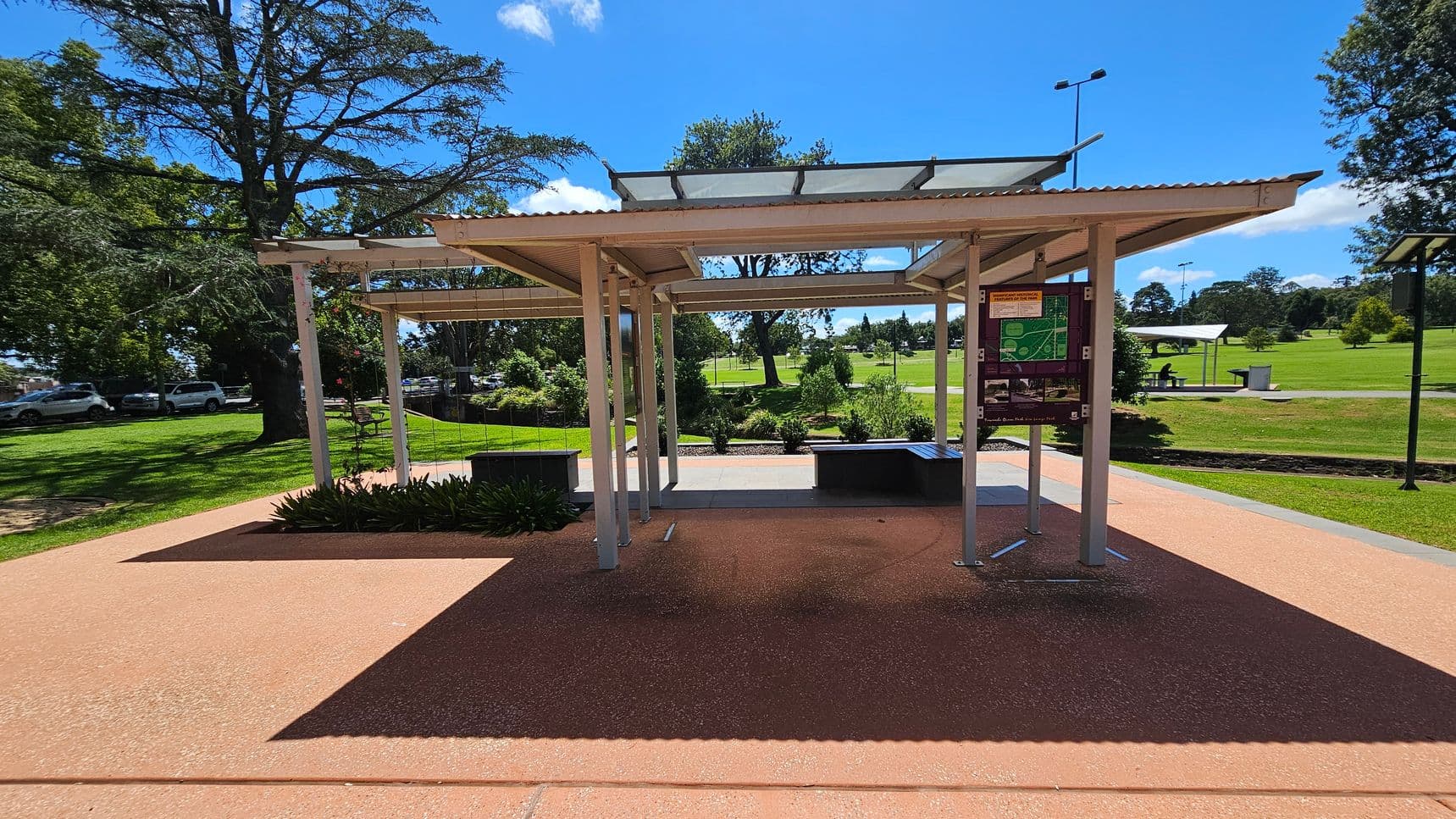 A park scene with a covered shelter in the centre, featuring a corrugated roof and transparent panels. To the right, there's a signboard with maps and information. Benches and planters are beneath the shelter. The background shows a grassy area with trees and a pathway. On the left, cars are parked under large trees. The ground is paved with a reddish-brown surface, and the sky is clear and blue.