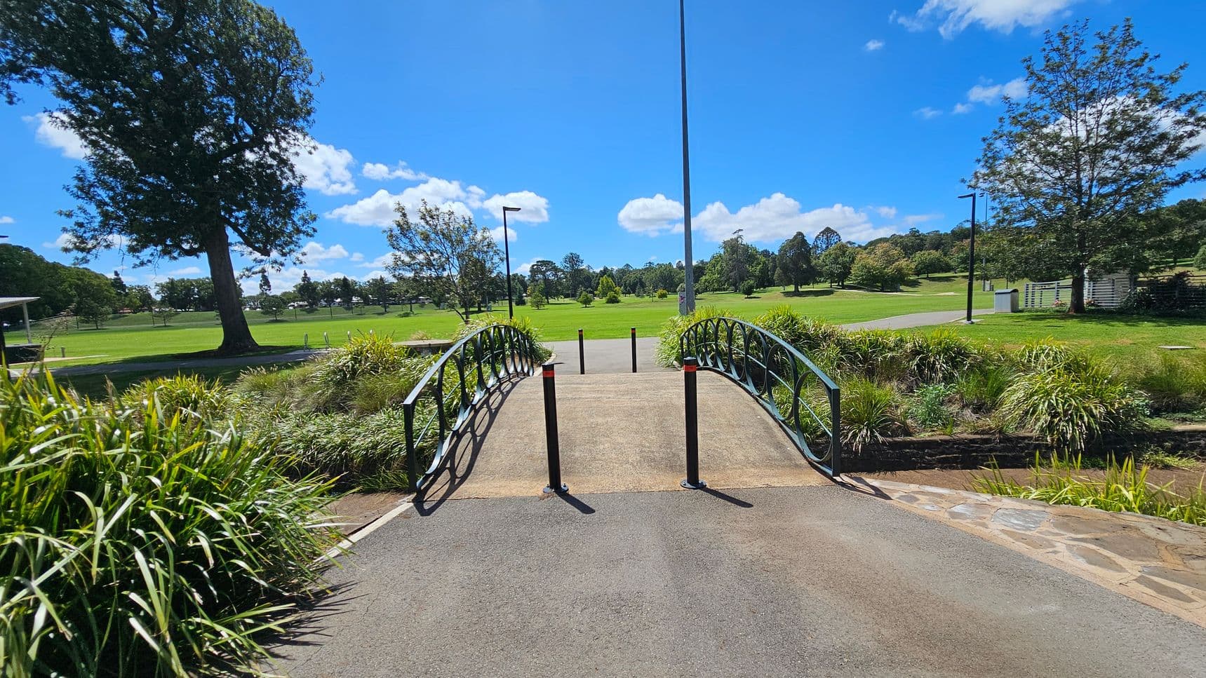 A small arched bridge with black railings crosses over a narrow stream. The bridge is flanked by tall grasses and shrubs. In the background, a vast green parkland stretches out, dotted with trees and a few lamp posts. A large tree stands prominently on the left, while a paved path leads into the distance. The sky is clear with a few clouds.