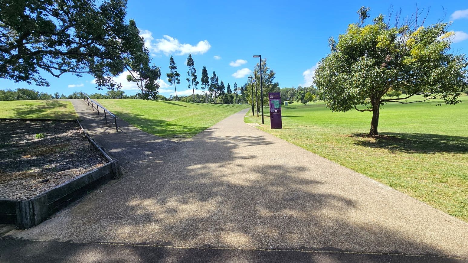A paved path runs through a park, flanked by grassy areas. On the left, a large tree with overhanging branches casts shadows on the path. A wooden-edged, mulched area is adjacent to the path. On the right, a solitary tree stands on the grass. Further along the path, several tall, slender trees line the route, and a signpost is visible. The sky is clear with a few clouds.
