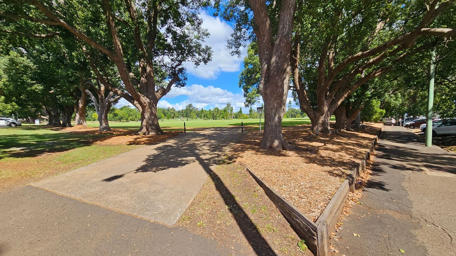 A pathway leads through a park with large, leafy trees on both sides. The ground is covered with fallen leaves and patches of grass. To the left, a car is parked near the trees. On the right, a paved footpath runs alongside a row of parked cars. In the background, an open grassy field stretches out, bordered by more trees. A wooden barrier lines the edge of the path on the right. Shadows of the trees are cast on the ground.