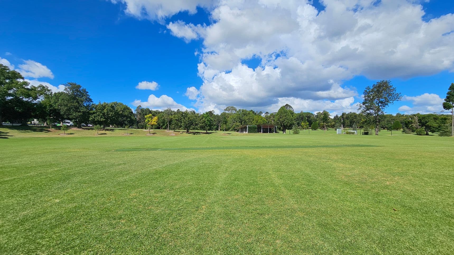A wide expanse of green grass covers the foreground, leading to a line of trees in the background. In the centre, there is a small structure, possibly a pavilion or shelter, surrounded by more trees. The sky is filled with large, fluffy clouds. The scene is open and spacious, with trees scattered throughout the area.