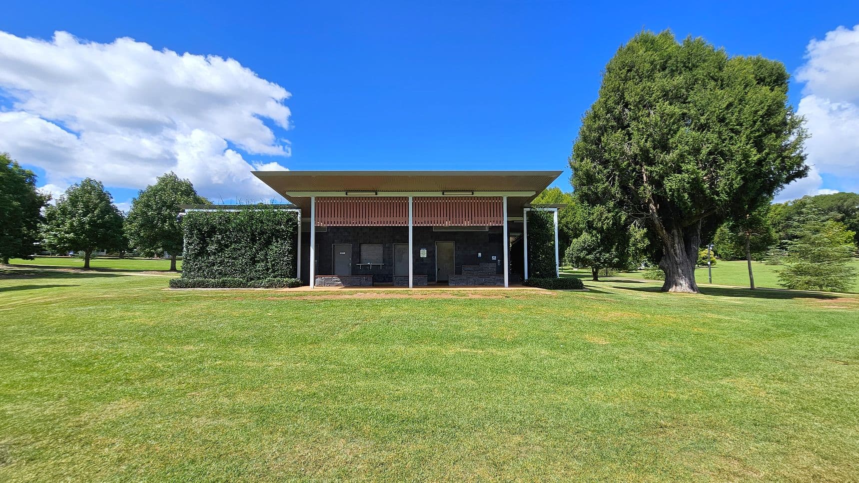 A small, rectangular building with a flat roof is centred in a grassy park. The building has a facade with vertical slats and is flanked by green hedges. On the left, several trees are visible in the background. On the right, a large, dense tree stands prominently. The foreground features a well-maintained lawn, and the sky is bright blue with scattered clouds.