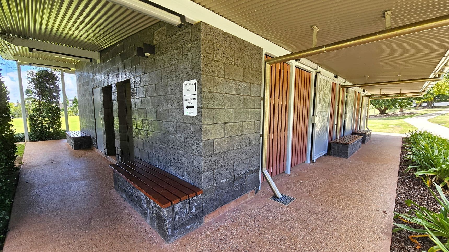 A public toilet facility with a corrugated metal roof. The left side has a dark brick wall with a sign indicating directions for event toilets and public toilets. Wooden benches are attached to the wall. The right side features wooden slats and metal doors. The floor is a textured concrete surface. In the background, there is a grassy area with trees and a pathway. Bushes line the right side of the walkway.