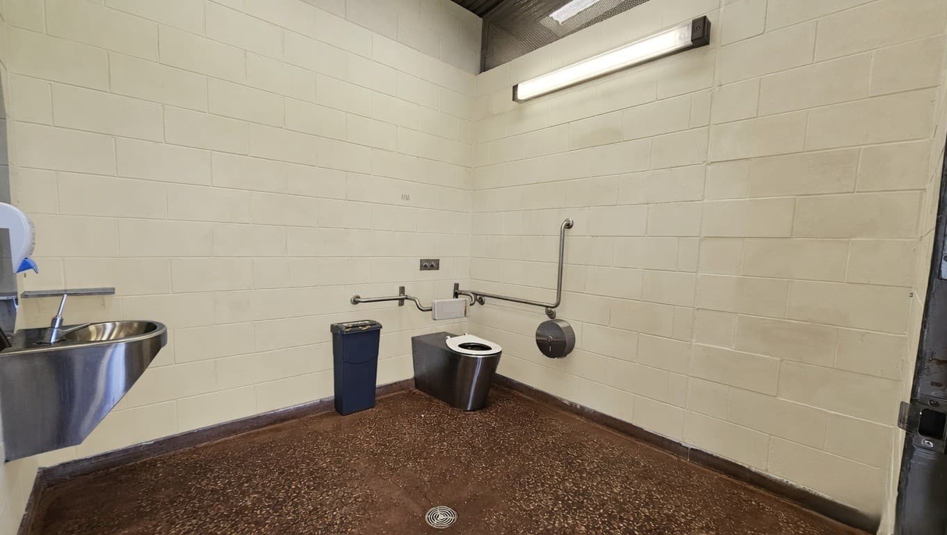 A public restroom with a stainless steel sink on the left, mounted on a light-coloured brick wall. In the centre, there is a metal toilet with a black seat, accompanied by a blue waste bin to its left. The walls have metal handrails, and a large toilet paper dispenser is mounted on the right. The floor is speckled brown, and a drain is visible. A fluorescent light fixture is mounted on the wall above the toilet.