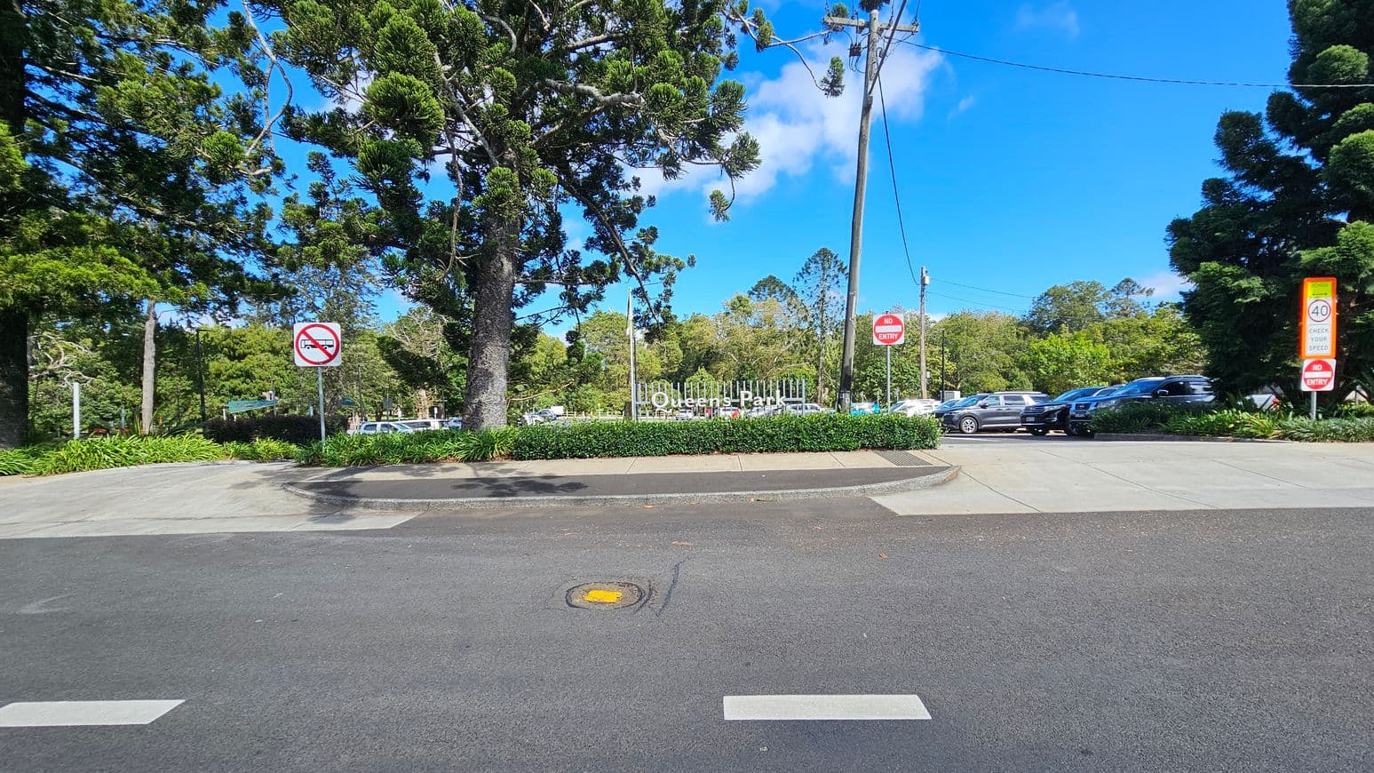 A road with a pedestrian crossing in the foreground. In the middle, a sign reads "Queens Park" behind a hedge. To the left, large trees and a "No Right Turn" sign. On the right, a parking area with several cars, a "No Entry" sign, and a speed limit sign indicating 40 km/h. The background features more trees and a clear blue sky.