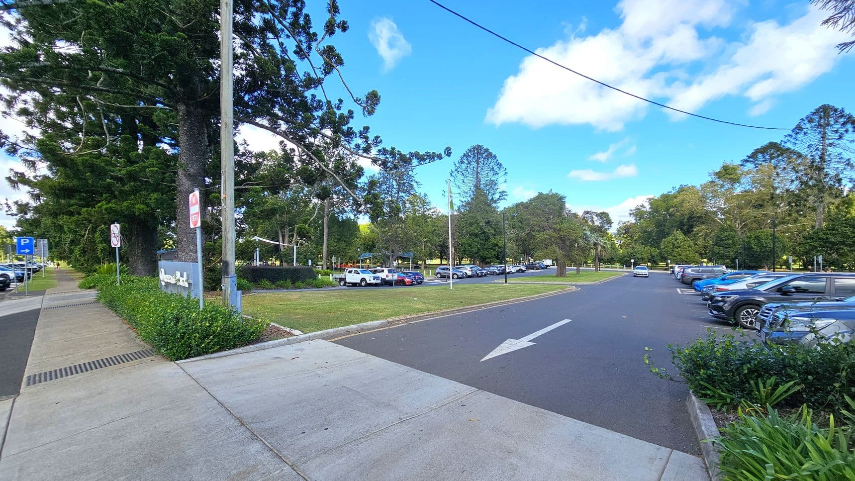 A paved footpath runs along the left side, bordered by a grassy area with large trees and a few signs. On the right, a car park is filled with vehicles, including a white truck and several blue cars. The road has a white arrow indicating direction. In the background, more trees and a park-like setting are visible. The sky is partly cloudy.