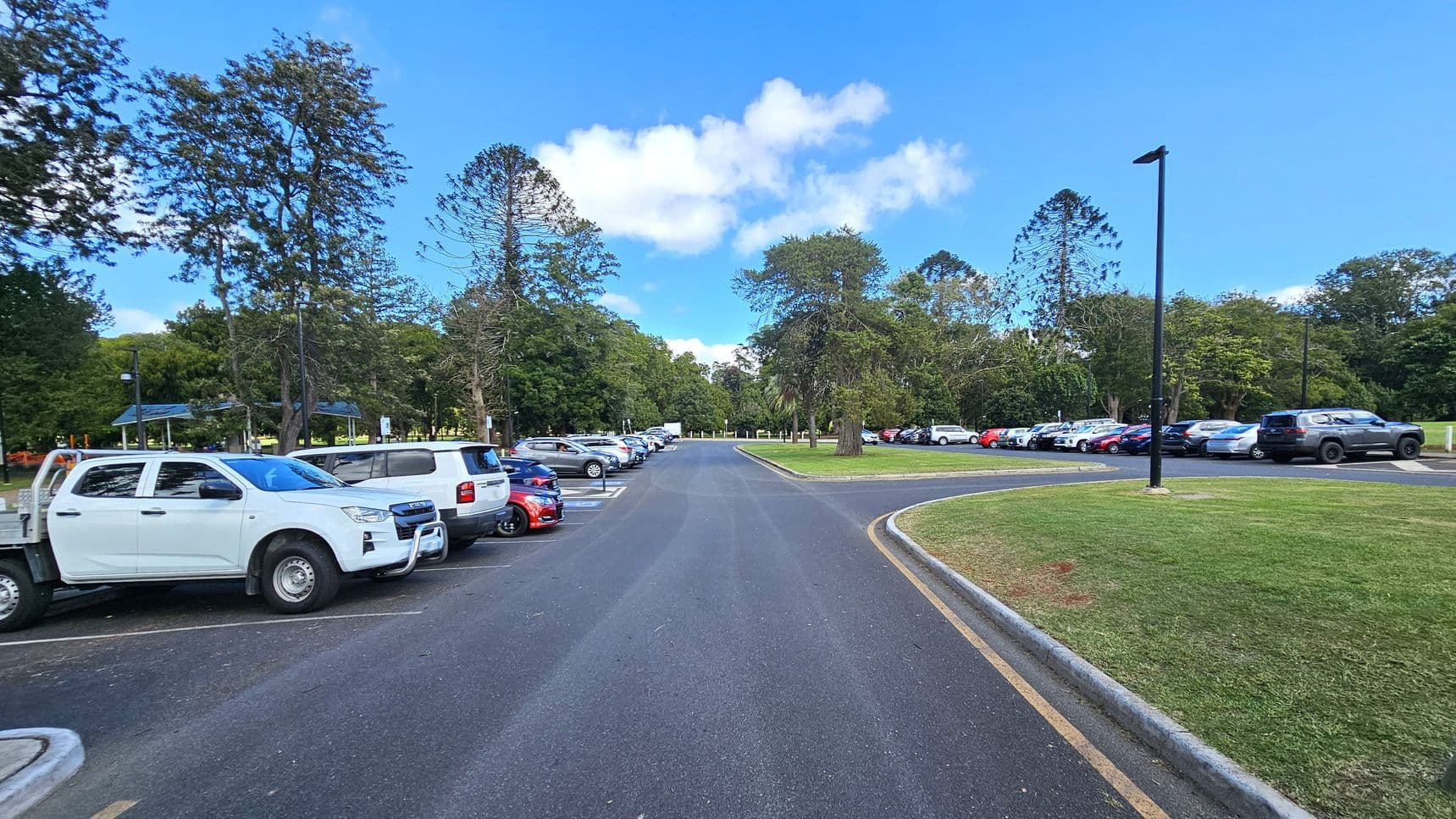 A car park with vehicles lined up on both sides of a central asphalt road. On the left, there are white and blue cars parked near tall trees and a small shelter. On the right, more cars are parked alongside a grassy area with a few trees. The background features dense greenery and tall trees. The sky is clear with a few clouds.