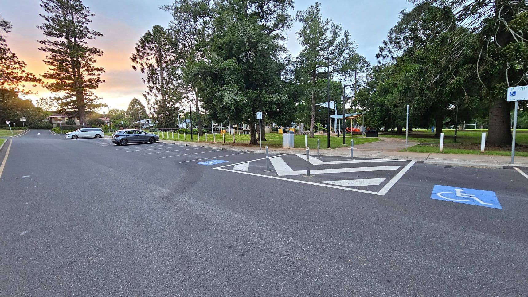 A car park with marked spaces, including a disabled parking spot in the foreground. On the left, two cars are parked near tall trees. The right side features a grassy area with more trees and a playground with a slide and swings. A path runs through the grass, and there are several bollards and signs. The background shows a few buildings and more trees under a colourful sky.