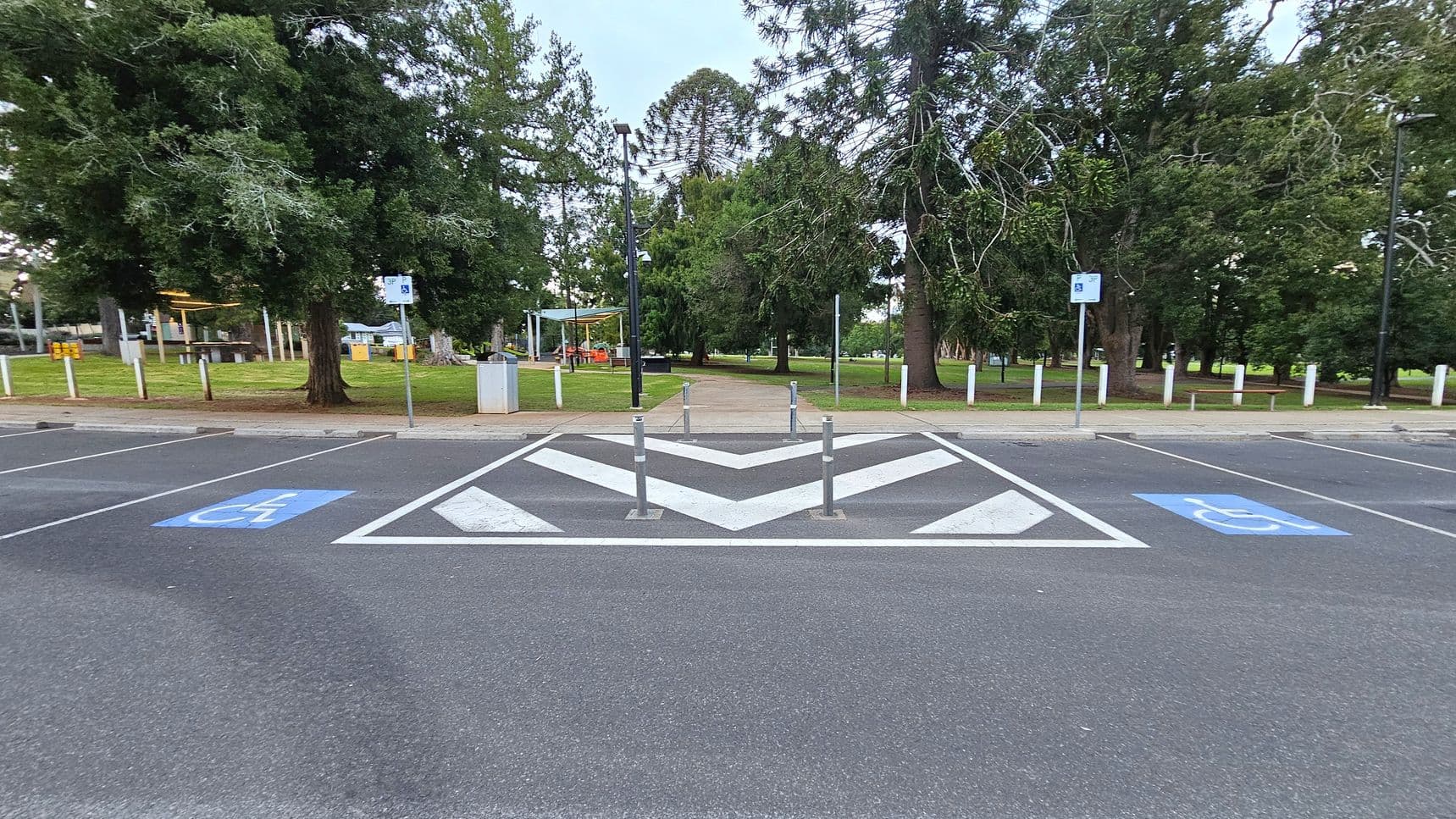 A paved parking area with two marked disabled parking spaces on either side. In the centre, there are white chevron markings and three bollards. Beyond the parking area, a path leads into a park with large trees and green grass. On the left, there is a covered picnic area with tables. Signs and lamp posts are visible along the path.