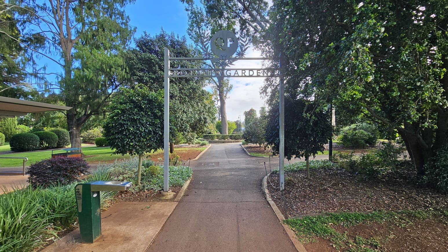 A pathway leads through a garden with a metal archway overhead, displaying the words "Botanic Gardens." On the left, there's a water refill station and a bench surrounded by greenery. The path is flanked by lush trees and shrubs, with a large tree visible in the background. The ground is a mix of concrete and soil, with well-maintained grass and plants lining the sides.
