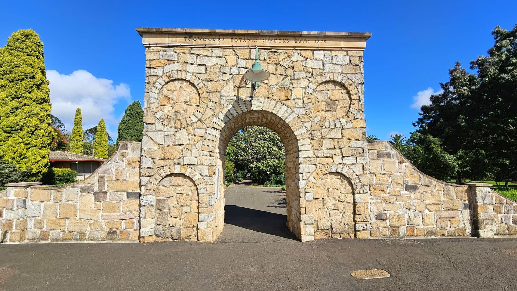 A stone archway with a central arched entrance and circular stone patterns on either side. The top of the archway has an inscription. A lamp hangs above the entrance. The path through the arch leads to a garden area with trees. On the left, there are tall, conical trees and a building with a tiled roof. On the right, more trees and greenery are visible. The ground is paved, and the sky is clear and blue.