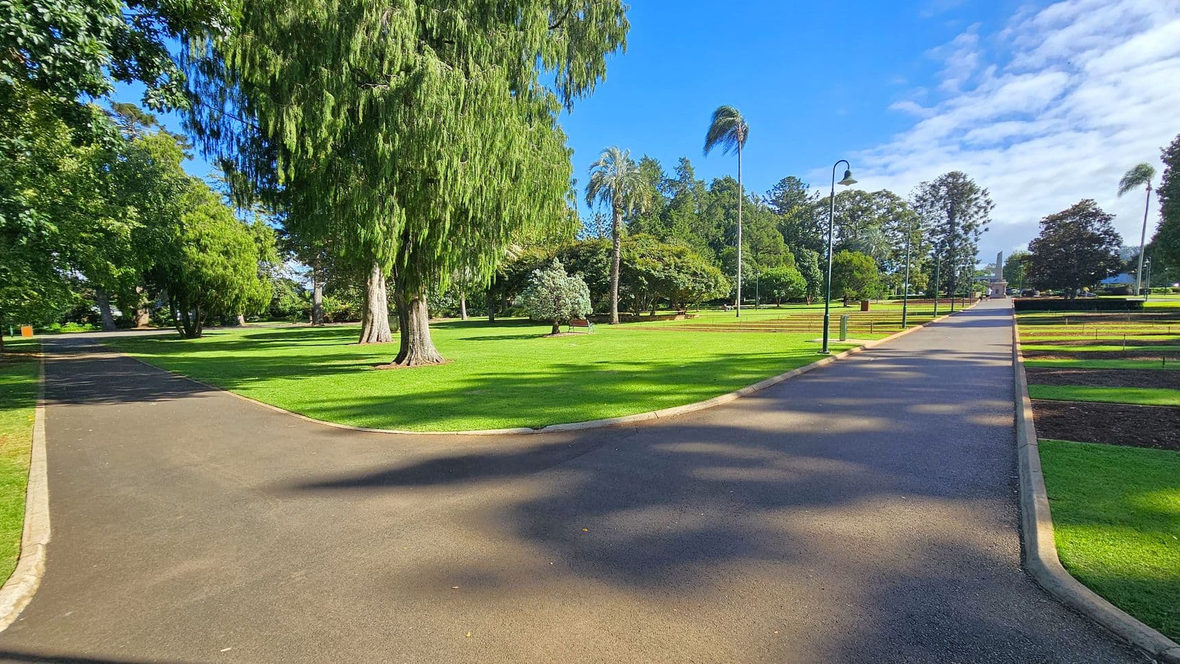 A park scene with a paved path splitting into two directions. On the left, the path curves through a shaded area with large, leafy trees. On the right, the path continues straight, lined with lampposts and bordered by neatly trimmed grass and flower beds. In the centre, a variety of trees, including a tall palm, stand on a well-maintained lawn. Benches are scattered throughout the area. The sky is clear with some clouds, casting shadows on the ground.
