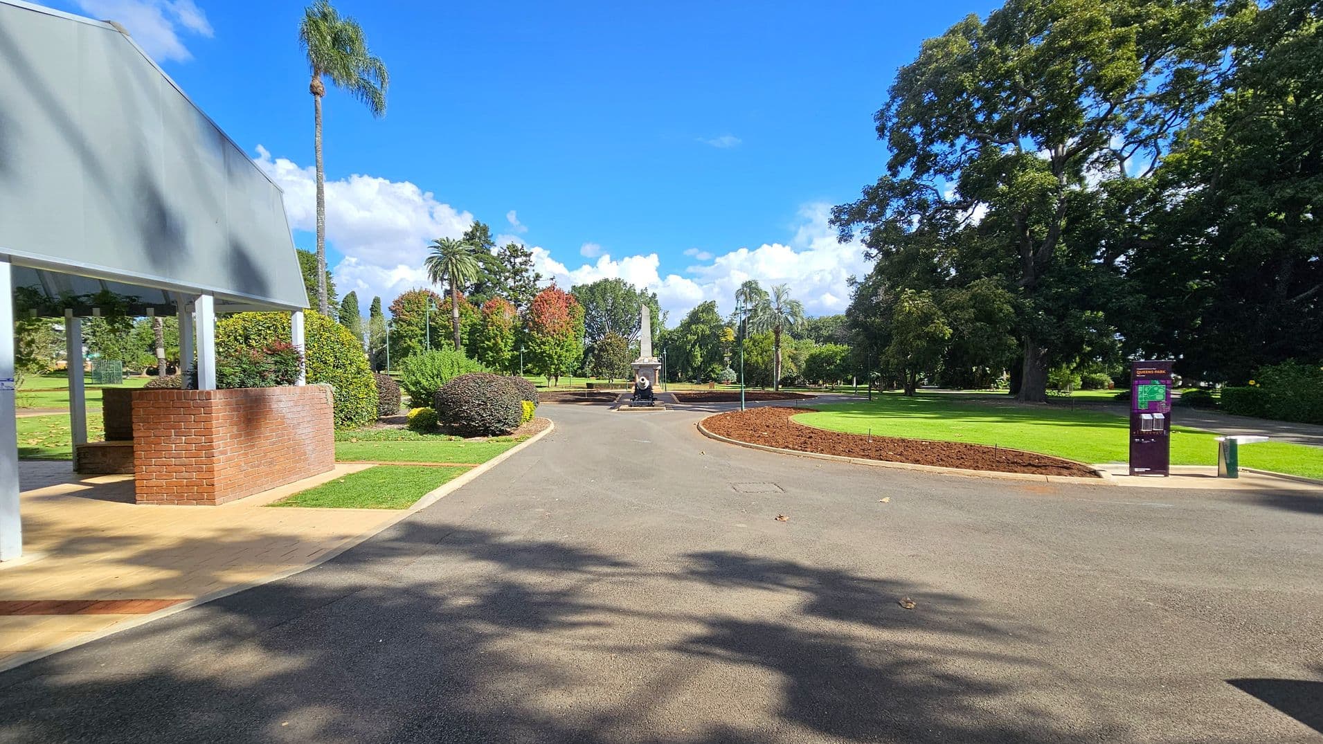 A paved pathway leads through a park with lush greenery. On the left, there's a structure with a brick base and a metal roof, surrounded by trimmed bushes. In the centre, a monument is visible in the distance. On the right, a large tree provides shade, and a signpost stands near a grassy area. The path is bordered by well-maintained lawns and flowerbeds.