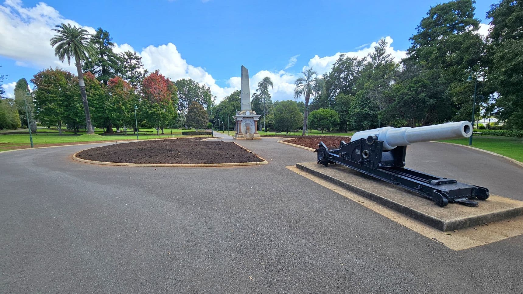 A paved pathway curves through a park, with a large cannon on a concrete platform to the right. In the centre, a tall obelisk stands on a decorative base. Surrounding the path are landscaped garden beds with dark soil. The background features lush green trees, including a prominent palm tree on the left. The sky is clear with some clouds.