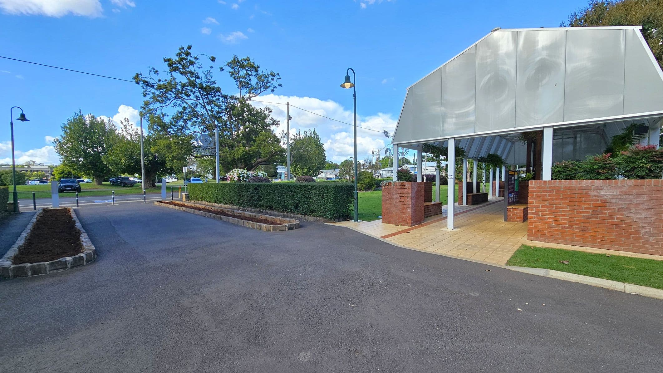 A paved area with a covered walkway on the right, featuring a structure with a metal roof and brick walls. The walkway is bordered by a small grassy area. On the left, there is a road lined with low stone borders and a hedge. Several street lamps are visible, and trees are in the background, along with parked cars. The sky is clear with a few clouds.