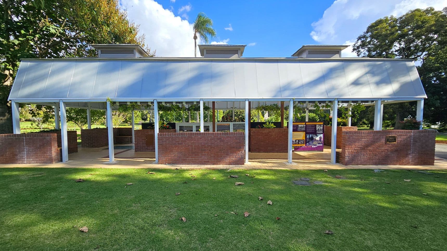 A structure with a sloped metal roof and three vented sections on top. The building has open sides supported by metal columns and is partially enclosed with red brick walls. Inside, there are informational panels and greenery. The foreground features a grassy area with scattered leaves, while the background includes trees and a clear blue sky.