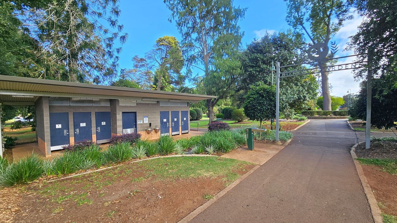 On the left, there is a public restroom facility with several doors, surrounded by green plants and a small garden area. In the middle, a paved path leads through a garden with neatly trimmed bushes and trees. On the right, an archway with the words "Botanic Gardens" marks the entrance to the garden area. The background features tall trees and more greenery, creating a lush, park-like setting.