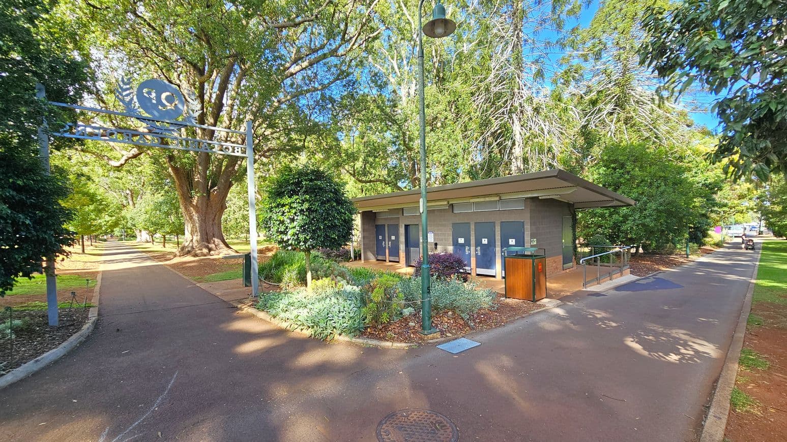 A park scene with a paved path running through the centre. On the left, a large metal archway with text leads to a tree-lined walkway. On the right, a small building with multiple doors, likely public toilets, is surrounded by greenery. A lamp post stands nearby, and a rubbish bin is positioned next to the building. Tall trees with dense foliage are in the background, providing shade. The ground is covered with a mix of pavement and mulch.
