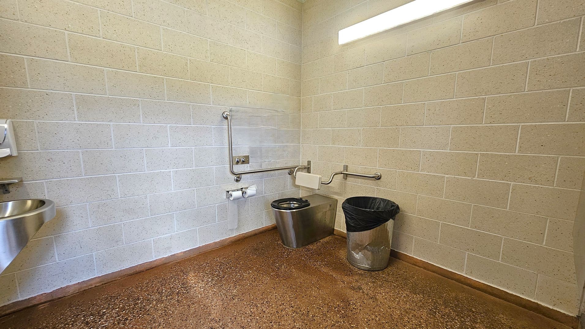 A public restroom with beige brick walls and a speckled brown floor. On the left, a stainless steel sink is mounted on the wall. In the centre, a stainless steel toilet with a black seat is equipped with grab bars on both sides. A toilet paper holder with two rolls is mounted on the wall. On the right, a metal bin with a black liner is placed on the floor. A fluorescent light fixture is mounted on the ceiling above the toilet area.