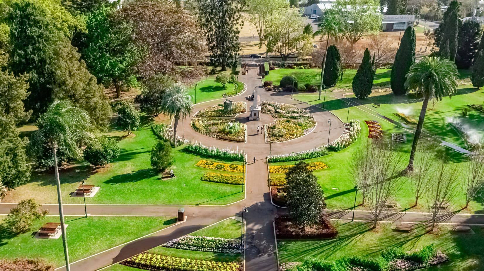Aerial view of a landscaped garden with a circular flowerbed in the centre, surrounded by pathways. The garden features vibrant flower arrangements and neatly trimmed lawns. Tall trees and palm trees are scattered throughout, with benches along the paths. Sprinklers are watering the grass on the right. In the background, there are more trees and a building labelled "Cobb+Co Museum". The scene is well-maintained and lush.