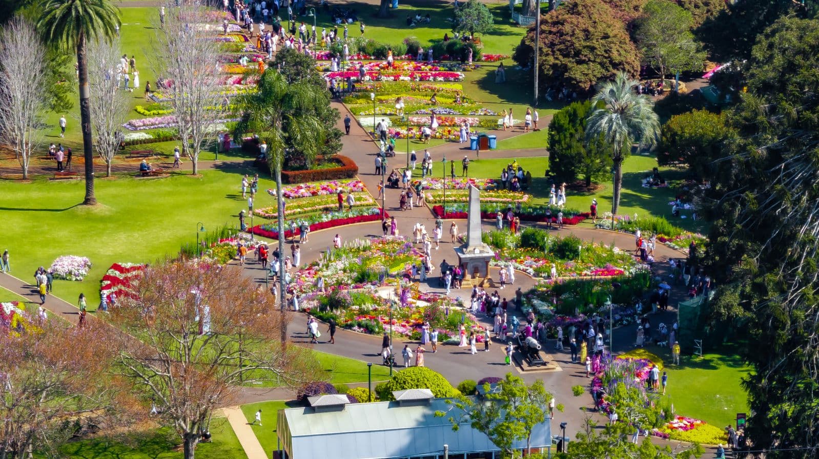 Aerial view of a vibrant garden with numerous flower beds arranged in geometric patterns. People are walking along the pathways, enjoying the colourful displays. Tall palm trees and other greenery are scattered throughout the area. In the centre, there's a monument surrounded by flowers. The foreground features a building with a light-coloured roof. The scene is bustling with visitors, indicating a lively atmosphere.