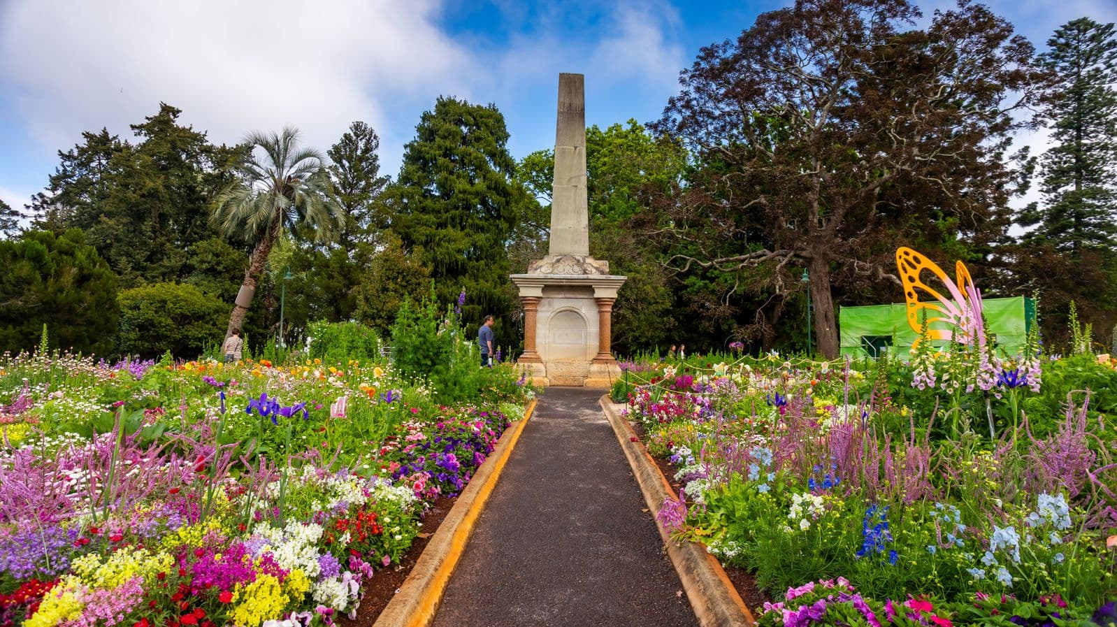 A vibrant garden with a central path leading to a tall stone obelisk. The path is bordered by colourful flowers in shades of purple, pink, yellow, and white. On the right, a large orange butterfly sculpture is visible. Tall trees, including a palm, form the background, creating a lush, green setting. The garden is well-maintained, with a mix of flowering plants and greenery.