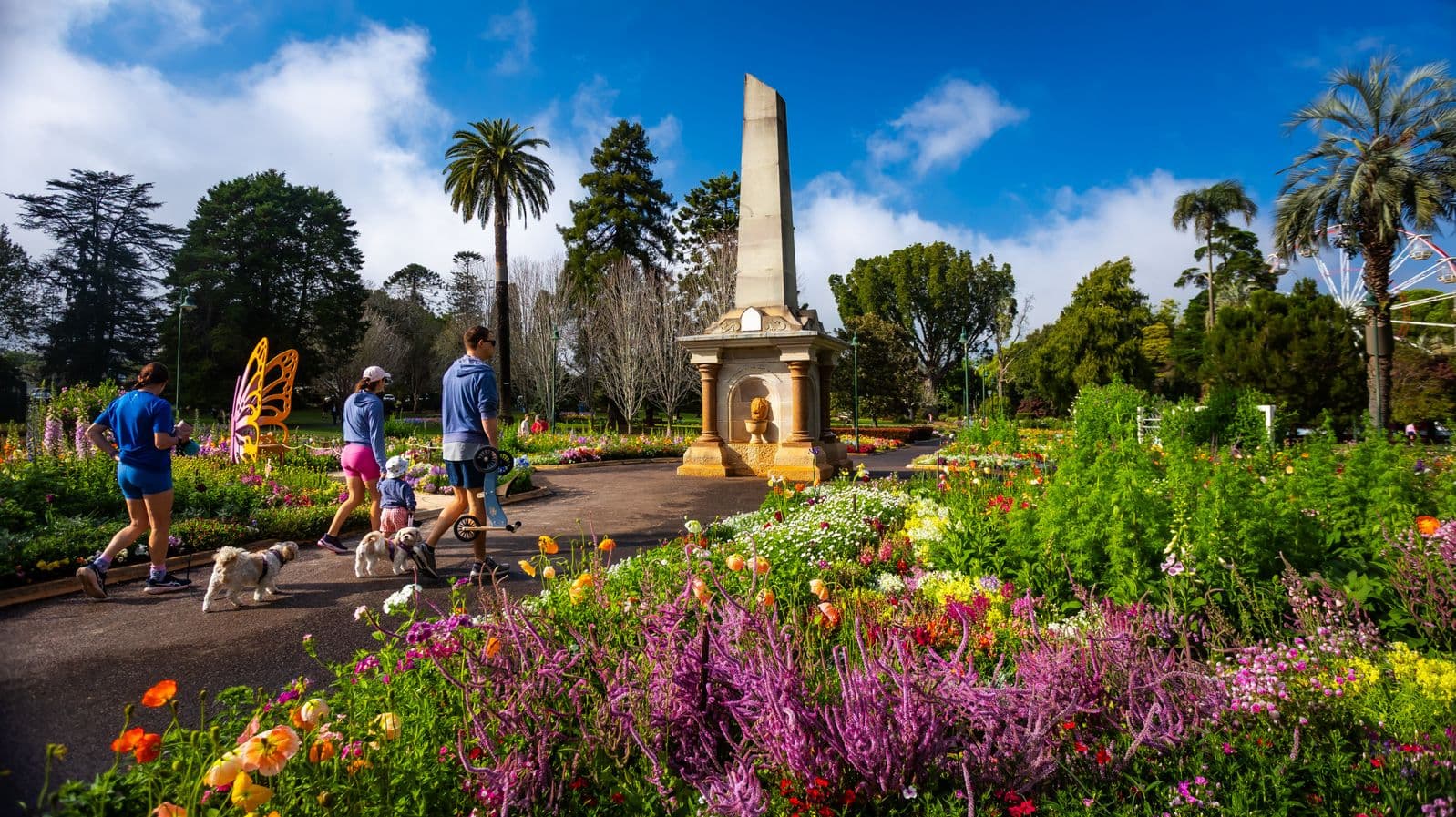 A vibrant garden scene with colourful flowers in the foreground, including purple, yellow, and orange blooms. In the centre, a tall obelisk monument stands surrounded by lush greenery. On the left, a group of people, including a child in a stroller and two dogs, walk along a path. A large butterfly sculpture is visible nearby. Tall trees and a Ferris wheel are in the background, under a bright blue sky with some clouds.