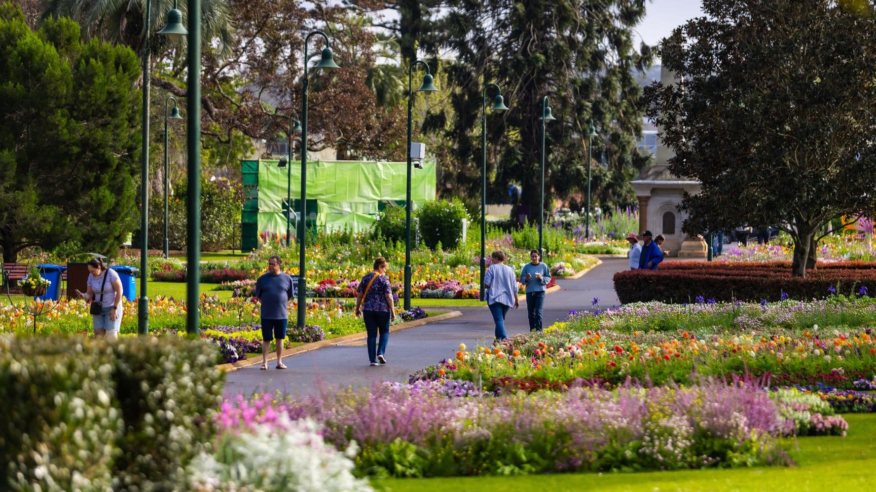 A vibrant garden scene with a paved path running through the centre. On both sides, there are colourful flower beds filled with various blooms. Several people are walking along the path, some stopping to admire the flowers. Tall green lamp posts line the path. In the background, there are large trees and a structure partially covered with green material. A hedge and more flowers are visible in the foreground.