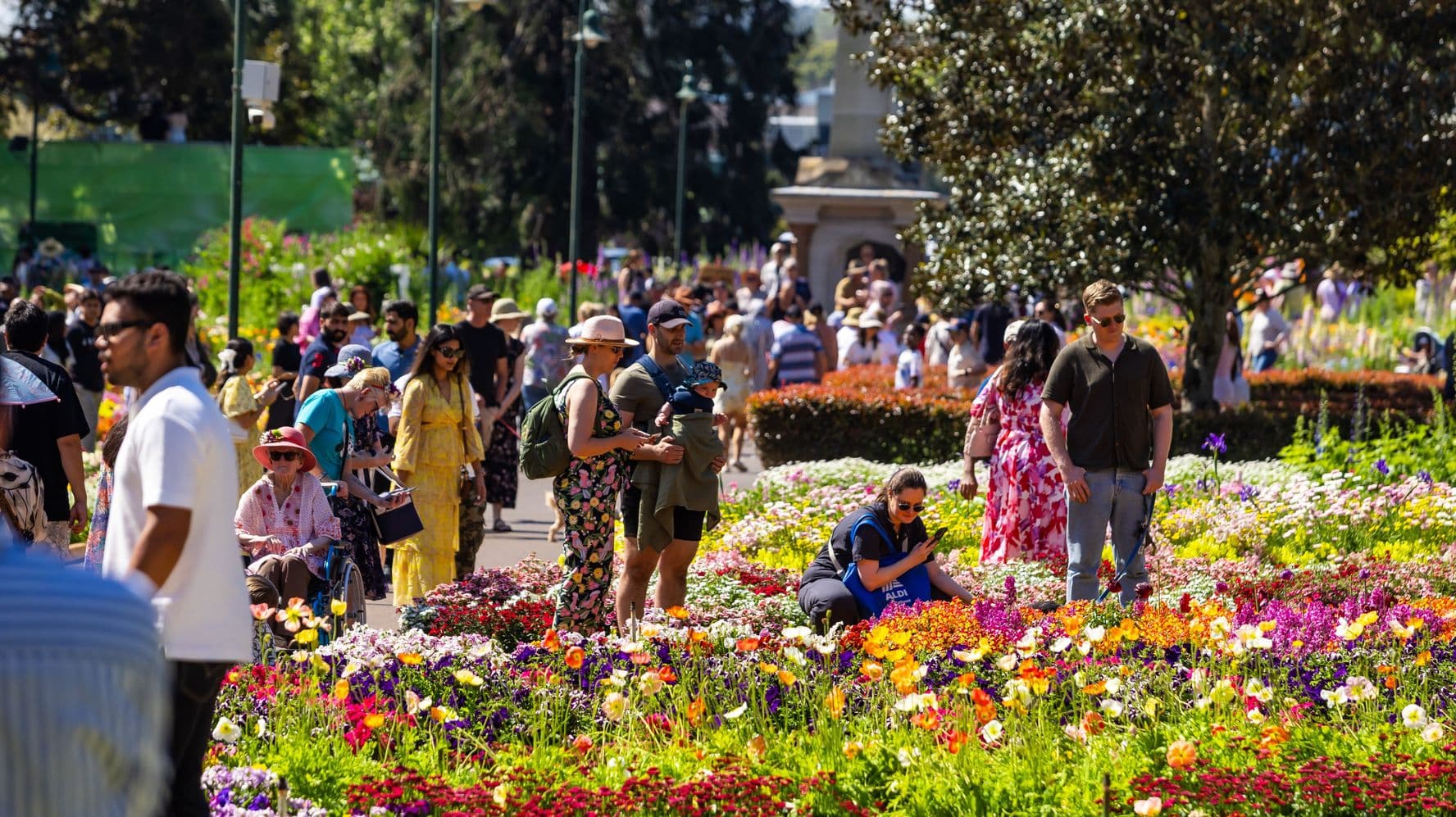 A bustling garden scene with numerous people walking and observing vibrant flower beds. On the left, a person in a wheelchair is accompanied by others, while a man holds a child. In the centre, a woman crouches to examine the flowers. On the right, a man in sunglasses stands near colourful blooms. The background features more people, trees, and a pathway lined with greenery. The atmosphere is lively and filled with diverse floral displays.