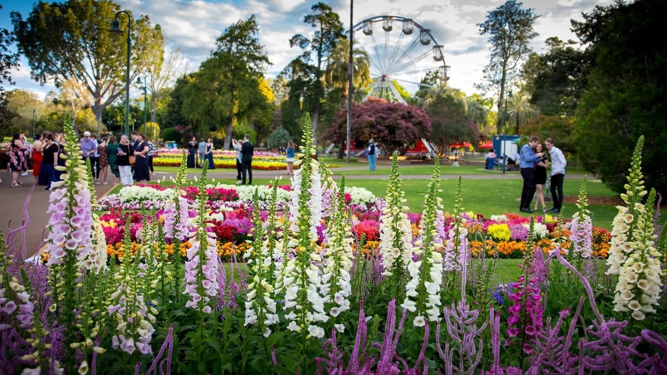 In the foreground, tall foxglove flowers in shades of purple and white stand prominently. Behind them, a variety of colourful flower beds are arranged in patterns. People are gathered on the left, some walking and others standing in groups. In the background, a Ferris wheel is visible among tall trees, adding a festive atmosphere to the park setting. The scene is lively with greenery and vibrant floral displays.