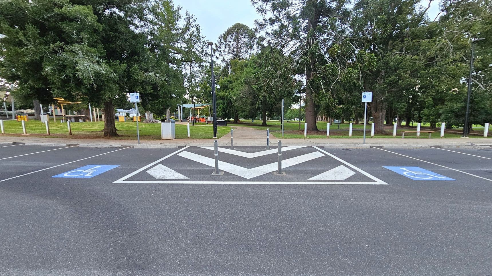 A parking area with two marked disabled parking spaces on either side. In the centre, there is a chevron pattern with three bollards. Beyond the parking area, a path leads into a park with large trees and a grassy area. On the left, there is a covered seating area with a picnic table. Signs and lamp posts are visible along the path. The ground is paved with asphalt.