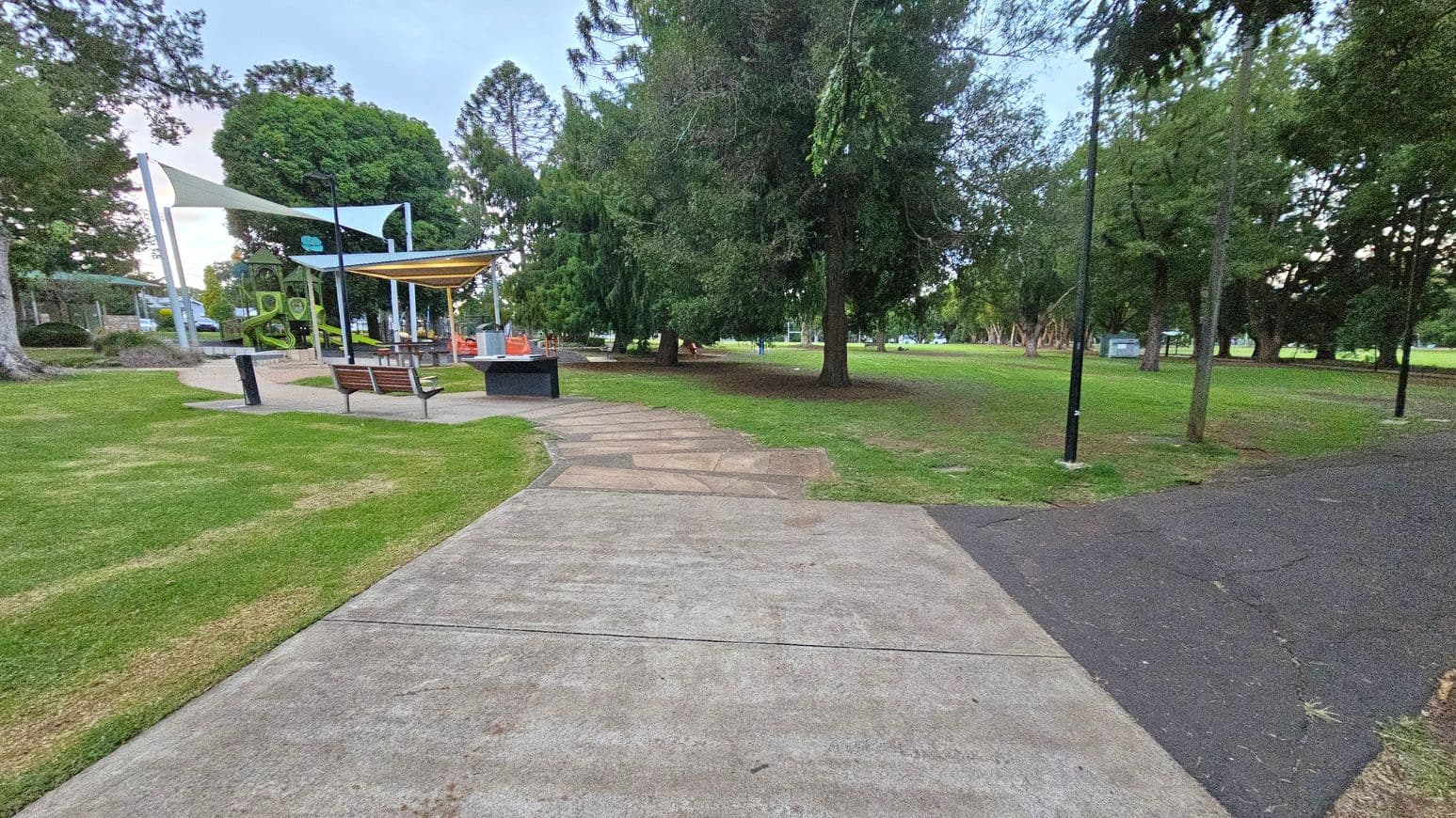 A park scene with a concrete path in the foreground leading to a playground on the left. The playground features green slides and is covered by shade sails. There are benches and a barbecue area nearby. On the right, a paved path runs alongside grassy areas and tall trees. Several lampposts line the paths, and more trees are visible in the background, creating a lush, green environment.