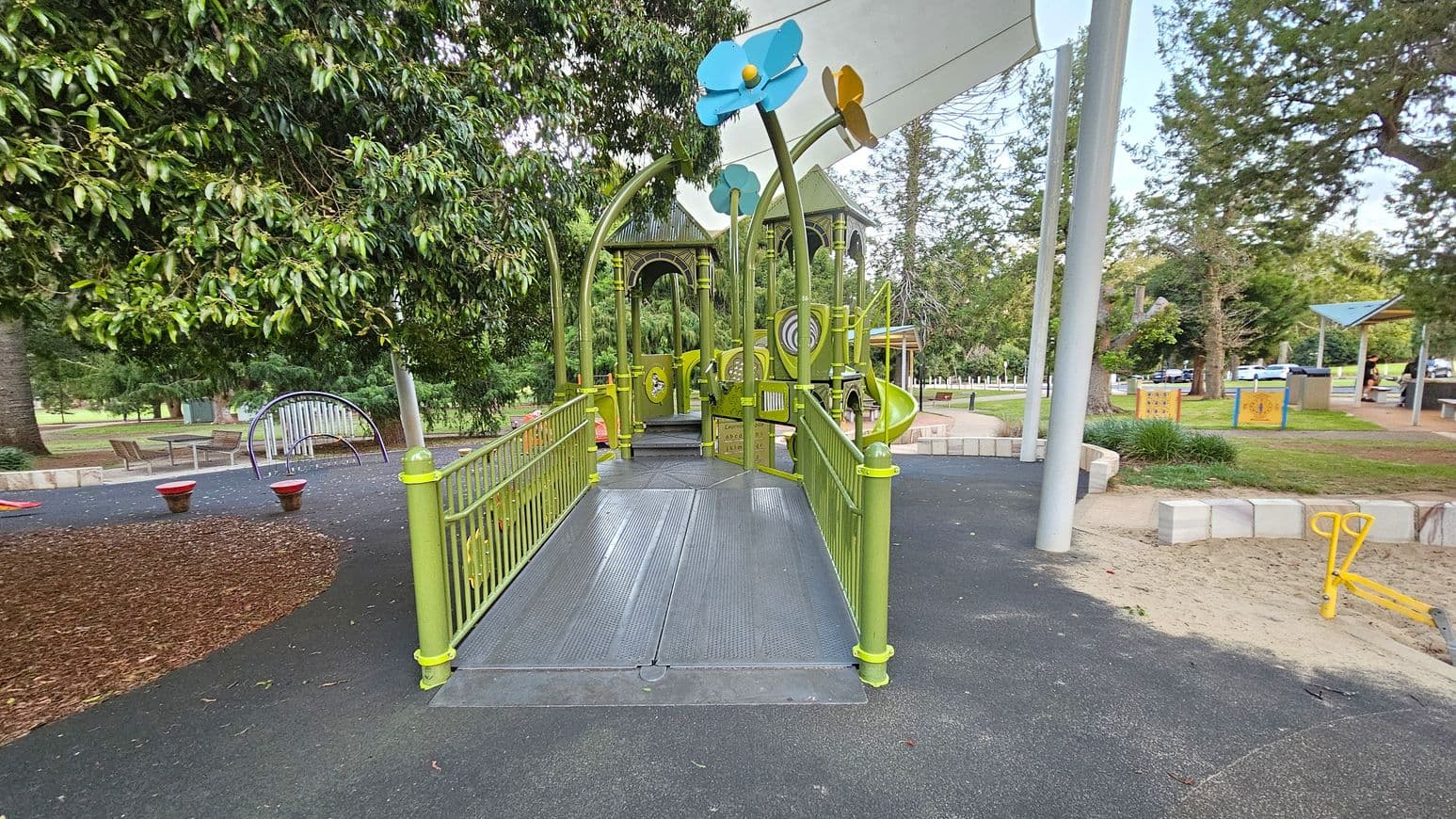 A playground with a green structure featuring a ramp leading to a play area with colourful flower decorations on top. On the left, there are trees and a curved climbing frame with benches nearby. The ground is covered with a mix of rubber and mulch. On the right, there's a sandy area with a yellow seesaw and a grassy section with picnic tables under a shelter. Trees and a car park are visible in the background.