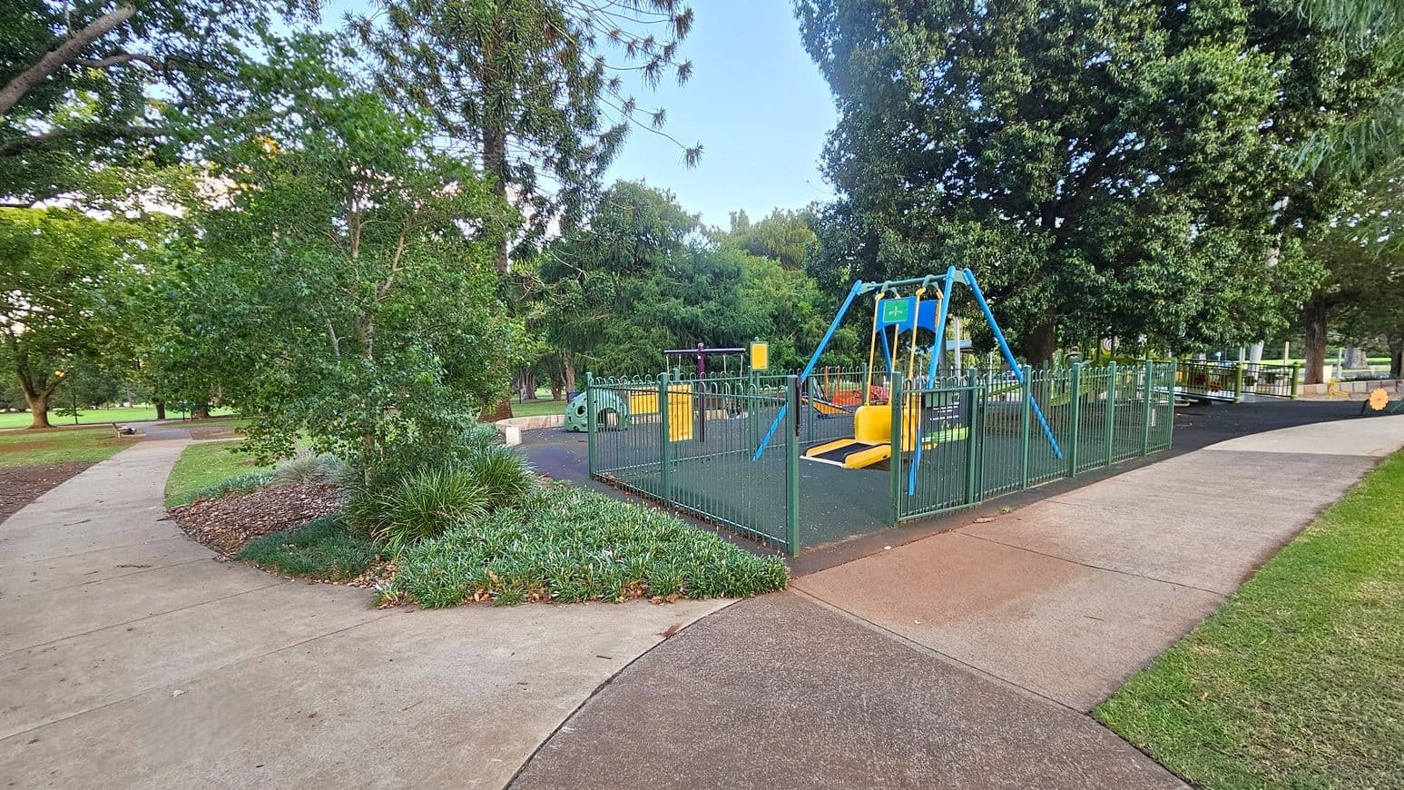 A playground is on the right, enclosed by a green metal fence. It features a blue and yellow swing set and other play equipment on a rubberised surface. To the left, a concrete path curves through a grassy area with trees and shrubs. The background is filled with more trees, creating a lush, green environment. The ground is a mix of grass and concrete paths.