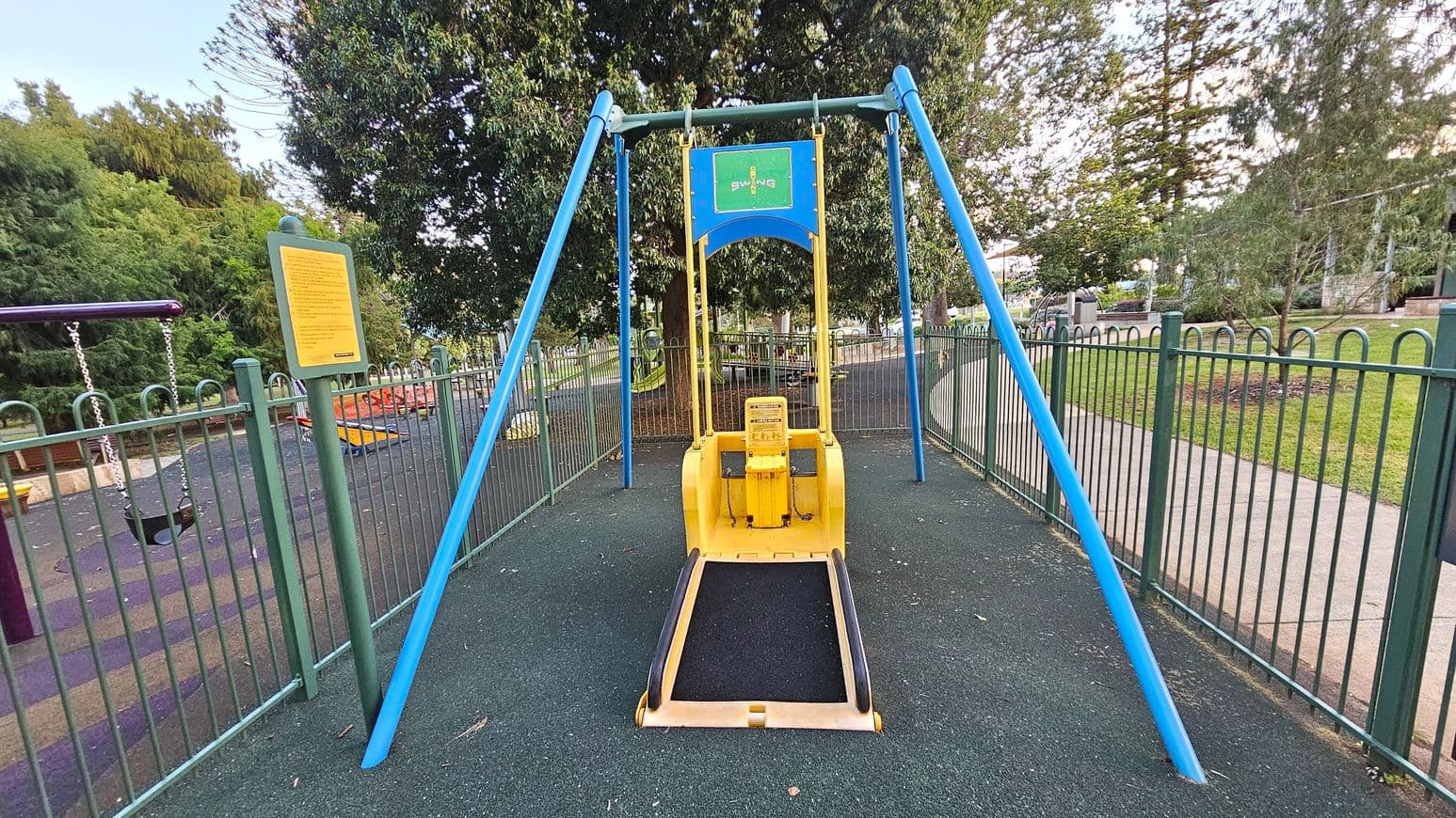 A playground scene with a large yellow swing designed for accessibility in the centre, supported by blue metal poles. To the left, there's a sign with text and a traditional swing set. The ground is covered with a rubber safety surface in shades of green and blue. In the background, there are trees and a grassy area, with a path running alongside the fenced playground.