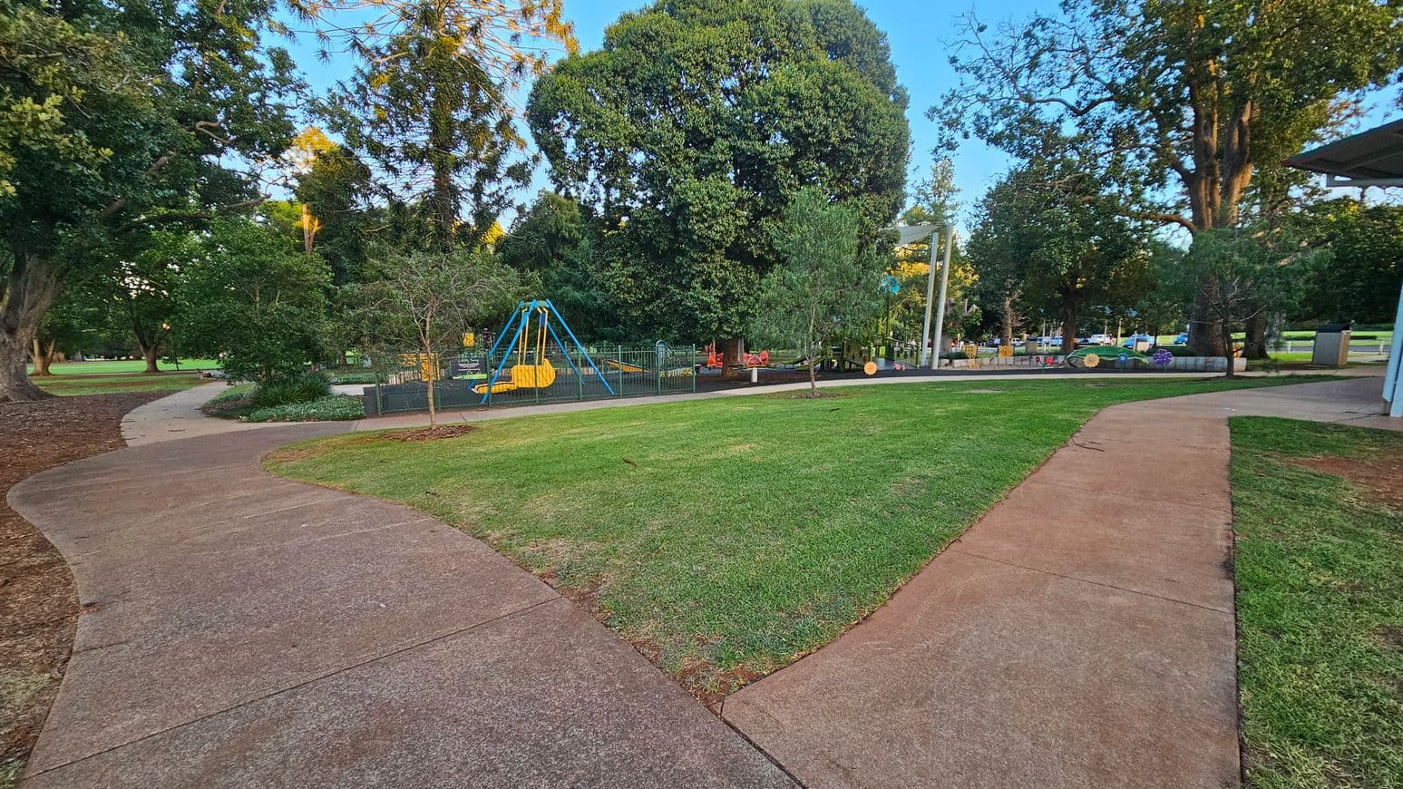 A park scene with a paved pathway curving through the foreground. On the left, there are large trees and a grassy area. In the middle, a fenced playground features a blue and yellow swing set and other play equipment. The right side shows more trees and a small structure with a roof. The background is filled with additional trees and greenery.
