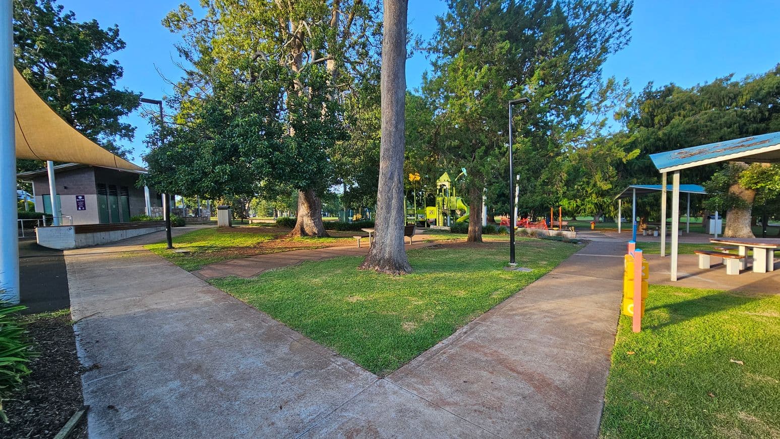 A park scene with a concrete path splitting into two directions. On the left, there's a building with a beige shade sail. In the centre, a large tree stands on a grassy area with benches. To the right, a playground with green and yellow equipment is visible among trees. A covered picnic area with tables is also on the right. The ground is a mix of grass and concrete paths, and there are several tall lampposts throughout the area.