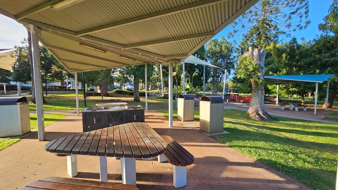 A park setting with a covered picnic area. In the foreground, there is a wooden picnic table on a concrete surface. The metal roof is supported by poles. To the right, there are two large rubbish bins and a tree with trimmed branches. In the background, more picnic tables are visible under blue shade sails, surrounded by green grass and trees. The area is well-lit, suggesting a sunny day.