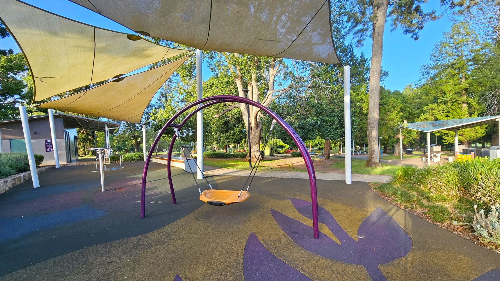 A playground with a large, circular swing in the centre, supported by a purple frame. The ground is covered with a colourful, patterned surface. Overhead, there are beige shade sails providing cover. To the left, a building with a slanted roof is visible, and to the right, there are picnic tables under a metal shelter. The background features tall trees and greenery, creating a lush park setting.