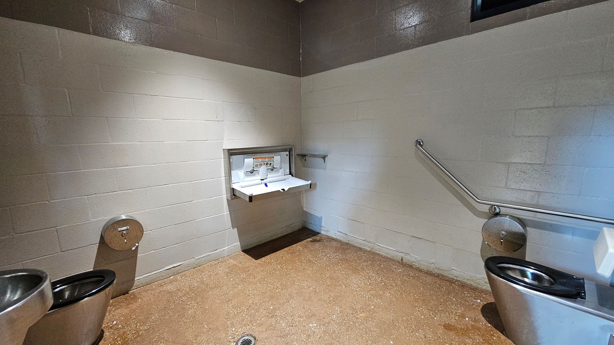 A public restroom with two stainless steel toilets on either side. In the centre, a baby changing station is mounted on the wall. The walls are made of painted cinder blocks, with a two-tone colour scheme. A metal grab bar is attached to the right wall. The floor is concrete with a drain in the middle. The ceiling is not visible.