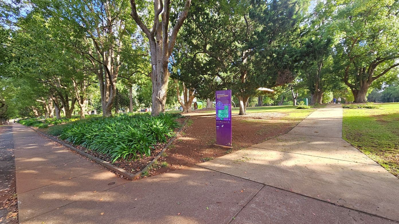 A park scene with a paved path on the left lined with tall trees and lush green foliage. In the centre, a purple sign stands on a patch of brown earth, surrounded by more trees. To the right, another path leads into the distance, bordered by grass and trees. The area is dappled with sunlight filtering through the leaves. In the background, a few people are walking along the path.