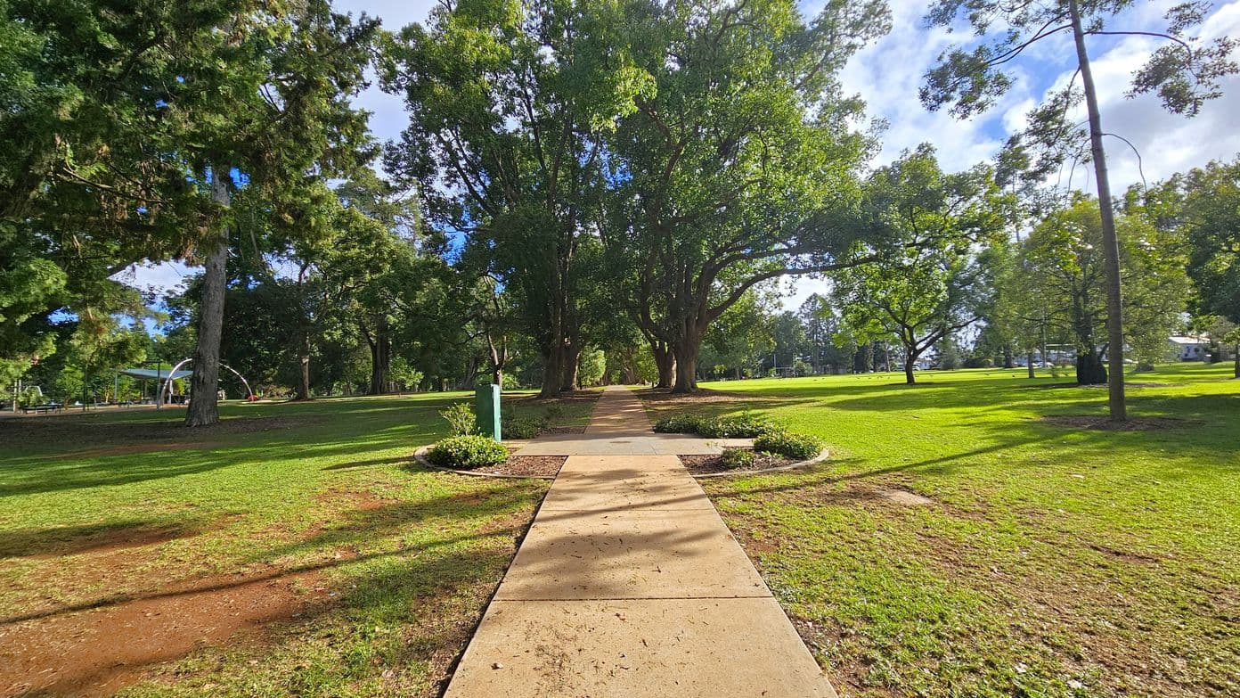 A paved path runs through the centre of a park, flanked by grass and trees. On the left, a playground structure is partially visible among the trees. The right side features more open grassy areas with scattered trees. In the background, large trees create a canopy over the path, casting shadows on the ground. The foreground shows a well-maintained lawn with some patches of dirt.