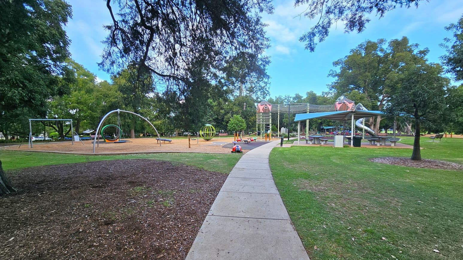 A park with a playground featuring various equipment. On the left, there are swings and a circular climbing structure. In the centre, a child is playing on the ground near a green spherical climbing frame. To the right, a covered picnic area with tables and a slide is visible. The area is surrounded by trees and grass, with a concrete path running through the middle.