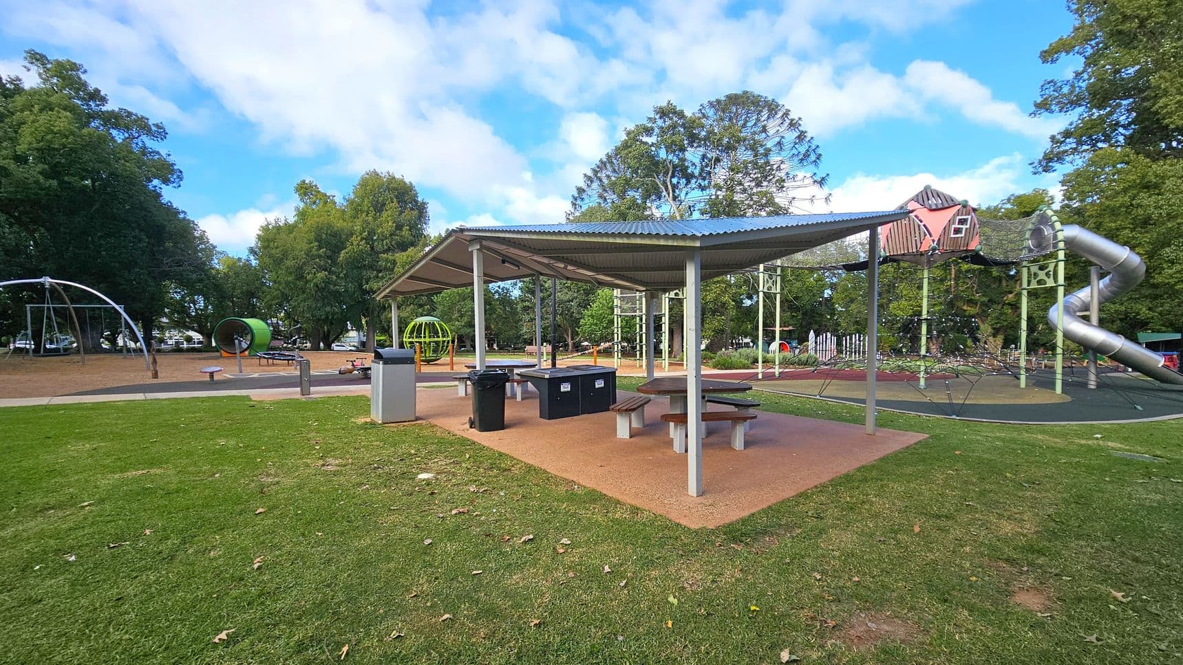 A playground with a covered picnic area in the centre, featuring tables and bins. To the left, there are swings and a large green circular play structure. On the right, a climbing frame with a red playhouse and a spiral slide is visible. The ground is a mix of grass and a rubberised surface. Tall trees surround the area, providing a natural backdrop.